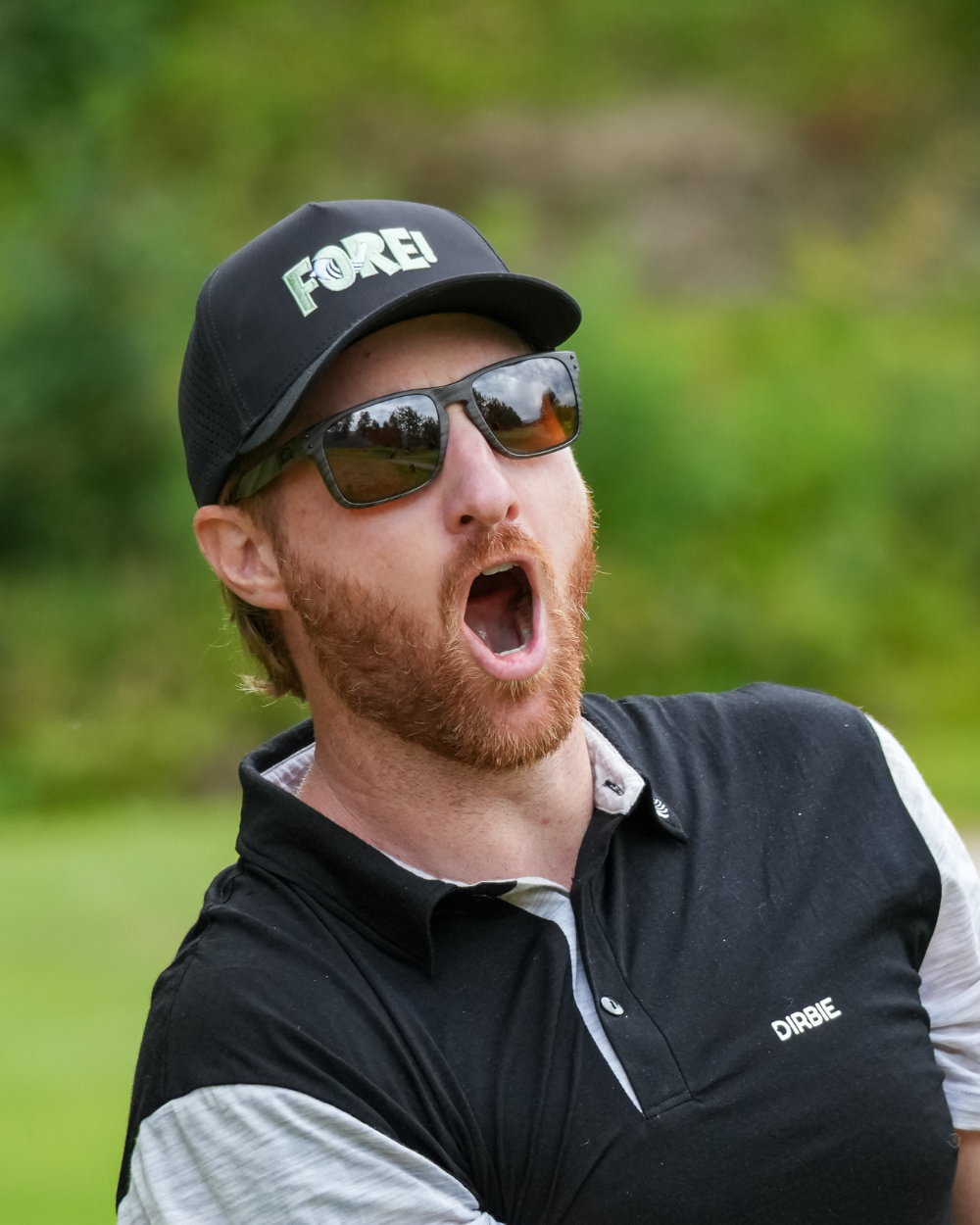 A man on a golf course wearing a black "Dirbie" FORE! Golf cap, dark sunglasses, and a black and white polo shirt, with his mouth open shouting "FORE".